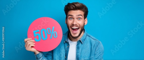A young man with a joyful expression is holding a large pink sign that shows fifty percent off. He stands in front of a bright blue background. The atmosphere feels fun and festive