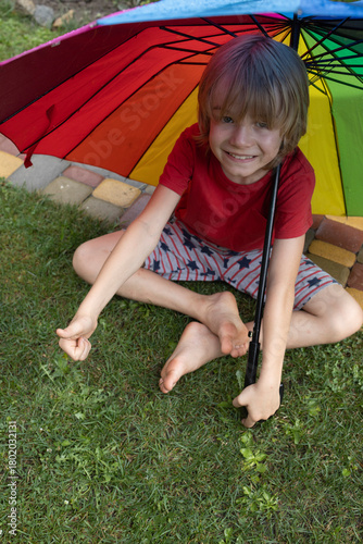 joyful barefoot child sits on the grass under a colorful umbrella during a summer rain. A carefree childhood, enjoying the freedom and coolness of the summer raindrops. Walking barefoot on wet grass