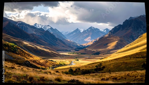 A wide, scenic view of a valley carved by a river, flanked by rolling hills and distant, snow-capped mountains, bathed in dramatic sunlight.
