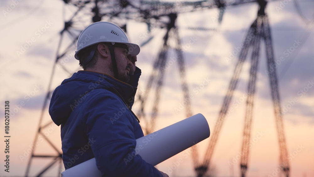 Obraz premium An electrician on the background of high towers of power plants looks at the project for the development of an electrical structure, the expansion of the electrical voltage of volts in the wires.