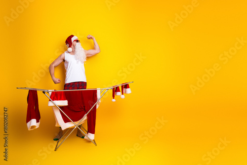 Fotografie Santa shows off a flexing arm beside a drying rack full of red outfits against a