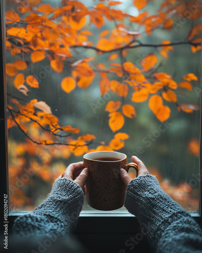a person holding a warm drink while looking out the window at orange leaves dri
