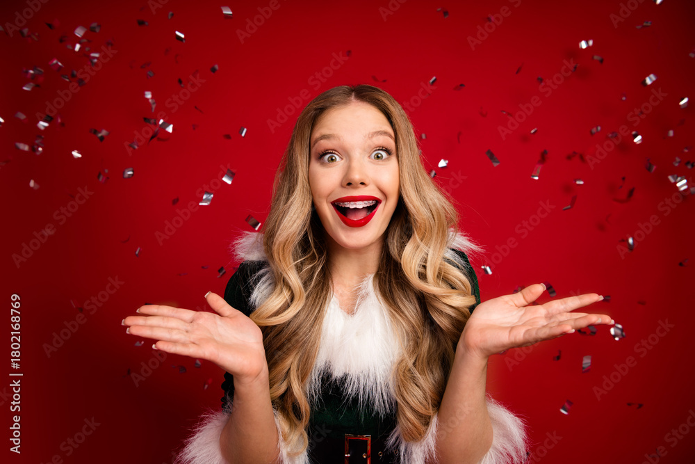 Naklejka premium Joyful girl in festive fur trimmed dress celebrates winter holiday with confetti against a red backdrop
