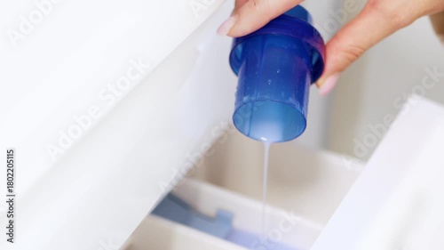 Hand pouring liquid laundry detergent or fabric softener into washing machine — clothes washing and home cleaning routine concept