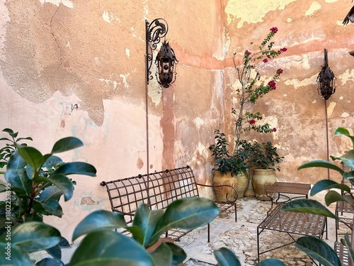 Mediterranean or vintage garden design and landscaping:Courtyard with iron furniture surrounded by terracotta pots planted with a climbing rose and green plants along a weathered wall with lanterns 