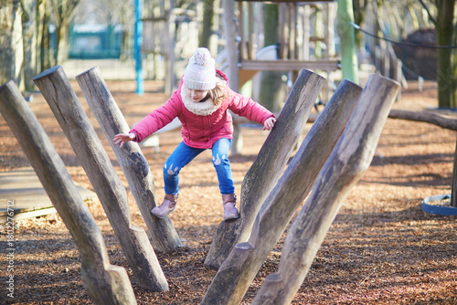 Bilde på lerret A young girl in warm winter clothing balancing and climbing on wooden playground