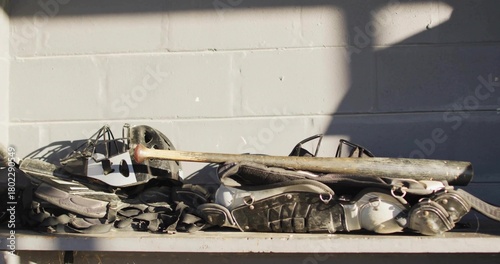 Sitting stacked catcher's gear on metal bench at dugout, chest pads, shin pads, masks, bat, straps