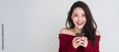 スマートフォンを両手で持って、うれしそうに笑っている若くて美人の女性、赤いオフショルダーニットを着ている