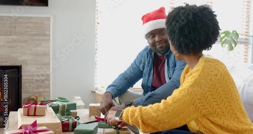 Picking up red ribbon spool, Diverse couple wrapping holiday gifts on coffee table for celebration