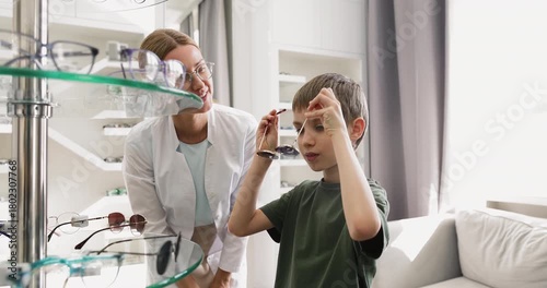 Young female optician helping to little boy with sunglasses selection