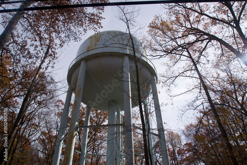 Fotografi Low-angle, dramatic view of a white, elevated municipal water tower (water tank) rising above the viewer, framed by a forest of trees with brown and orange autumn foliage