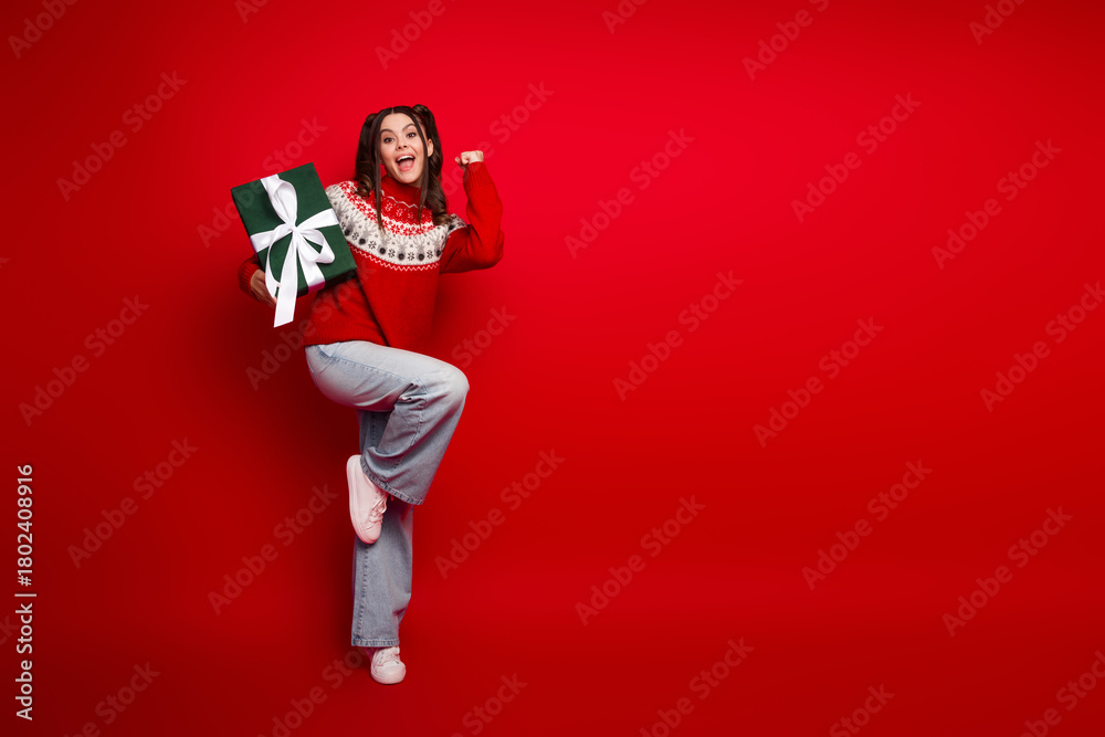Fototapeta premium Young woman in a festive red sweater holding a green gift against a solid red background celebrating Christmas joy and holiday season