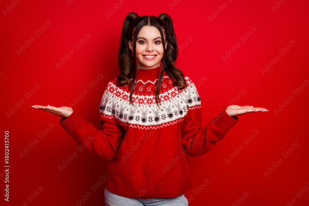 Fototapeta premium Young woman wearing a red Christmas sweater smiles against a solid red background with open hands for festive stock photo
