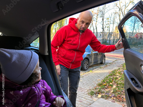 A parent leans toward the open car door to check on a child seated inside. The moment captures everyday parenting, attention to child safety, and preparing a child for a car ride