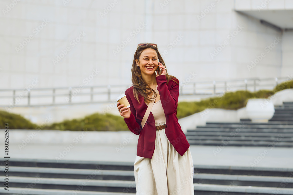 Fototapeta premium A stylish woman wearing a burgundy blazer and beige dress chats on her phone while holding a coffee cup in an urban outdoor setting. Bright, cheerful mood with a casual business vibe.