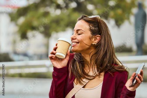 A stylish woman in a burgundy blazer smiles while sipping a takeaway coffee outdoors, holding a smartphone. Bright daytime urban scene conveys casual work, wellness, and contemporary lifestyle.