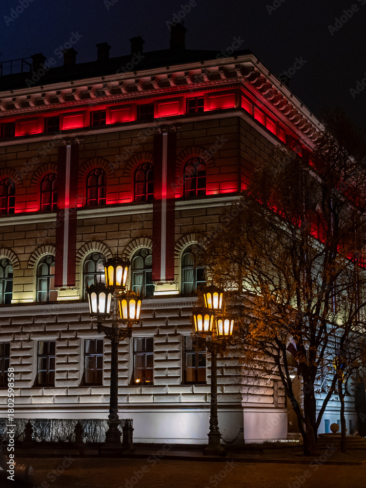 Obraz premium A historic building in Riga illuminated at night with striking red and warm white lights, accented by ornate street lamps and autumn trees, creating a dramatic and atmospheric city scene.