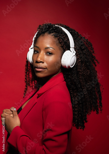 Hispanic woman with curly hair in red blazer wearing headphones listening music on red background