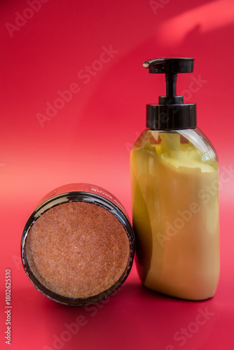 Beauty products showcased on a bright red backdrop, including an exfoliating scrub jar and a moisturizing lotion pump bottle, ideal for skincare routines