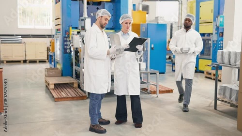 Three lab colleagues wearing protective coats and hairnets checking aluminum trays and discussing production quality. Team conducting inspection at food packaging factory. Industrial machinery.