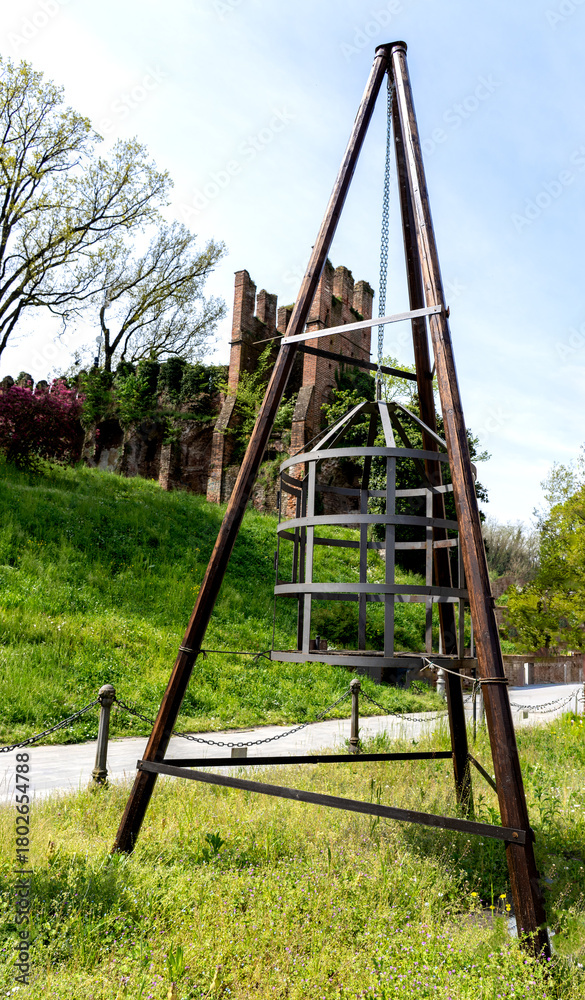 Fototapeta premium an ancient medieval torture device shaped like an iron cage for prisoners. In the background, you can see the remains of a medieval castle with characteristic battlements.