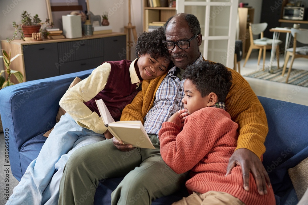 Obraz premium Senior Black man sitting on sofa reading book to two Black children, both kids leaning against him and listening attentively, cozy family moment in home living room setting