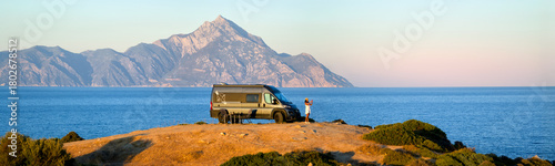 Woman enjoys the Aegean view beside a camper van with Mount Athos in the background. Ideal for: vanlife travel, outdoor brands, road-trip blogs, Greece tourism. Colours: blue, beige, silver, gold.