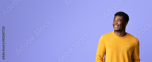 A man with a joyful expression stands against a purple wall. He is wearing a bright yellow sweater and appears happy, enjoying a light-hearted moment. The setting is bright and uplifting.