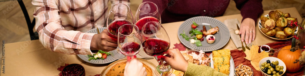 Obraz premium Header of diverse group of young and middle aged friends clinking wine glasses over Thanksgiving dinner table, visible hands and arms of Black man, Caucasian woman, sharing meal