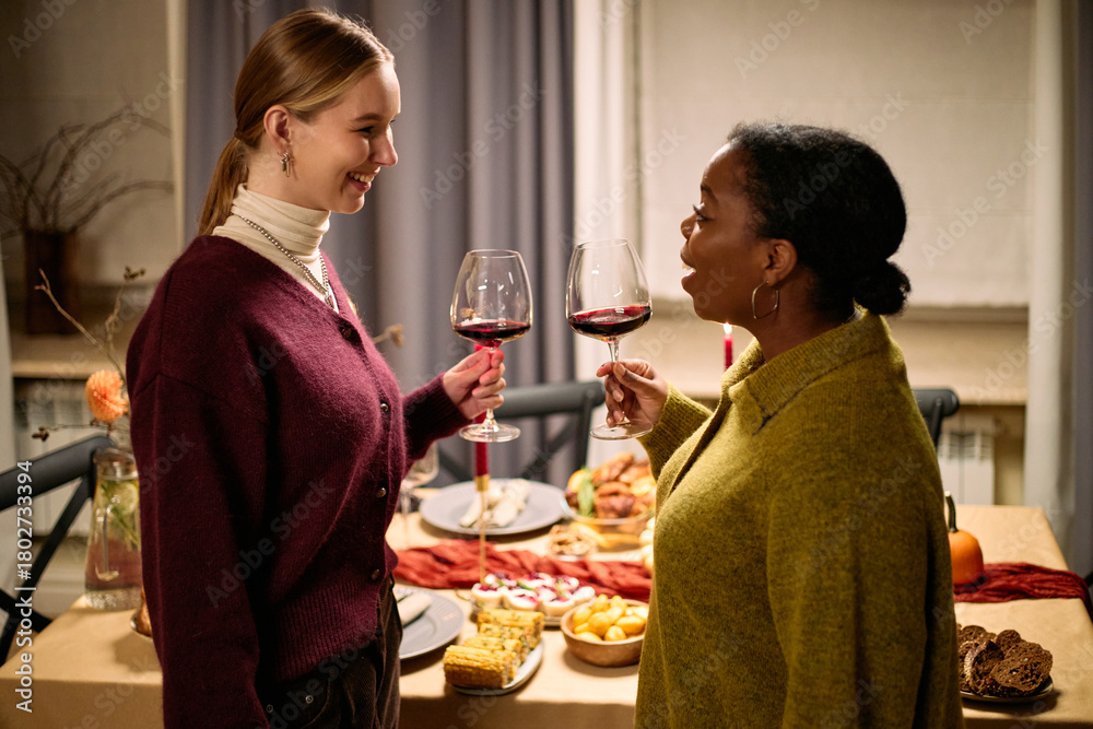 Fototapeta premium Young adult Caucasian woman and young adult Black woman standing near table holding wine glasses, smiling and talking during Thanksgiving dinner among friends, food visible in background