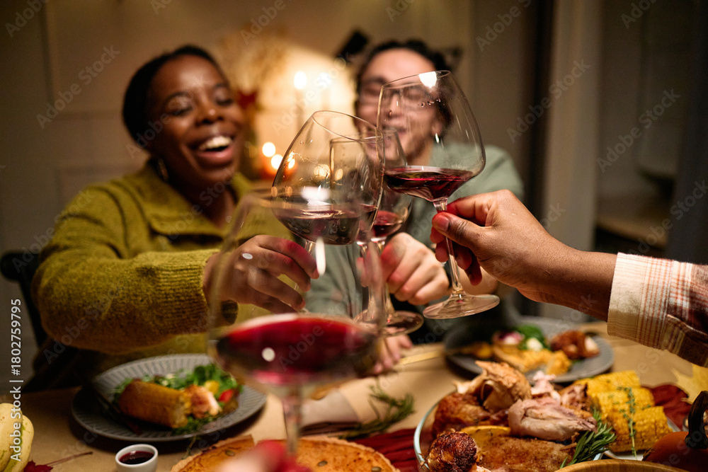 Obraz premium Diverse group of young people and middle aged friends clinking wine glasses while celebrating Thanksgiving dinner, plates with traditional holiday food on table, smiling and enjoying meal together