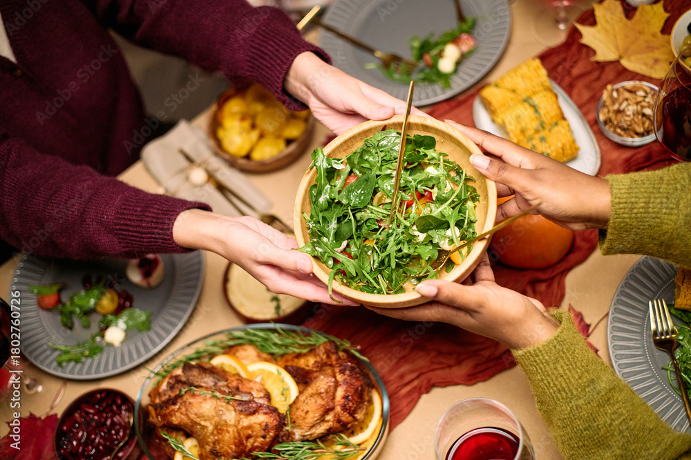 Obraz premium Diverse group of young adult and middle aged hands serving salad at Thanksgiving dinner table, sharing food among friends, visible plates with turkey, vegetables, and festive dishes