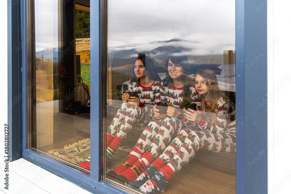 Fototapeta premium Family in pajamas enjoying cabin view indoors. Three girls wearing festive pajamas sit inside a cozy cabin, holding mugs and looking out at a scenic mountain landscape.