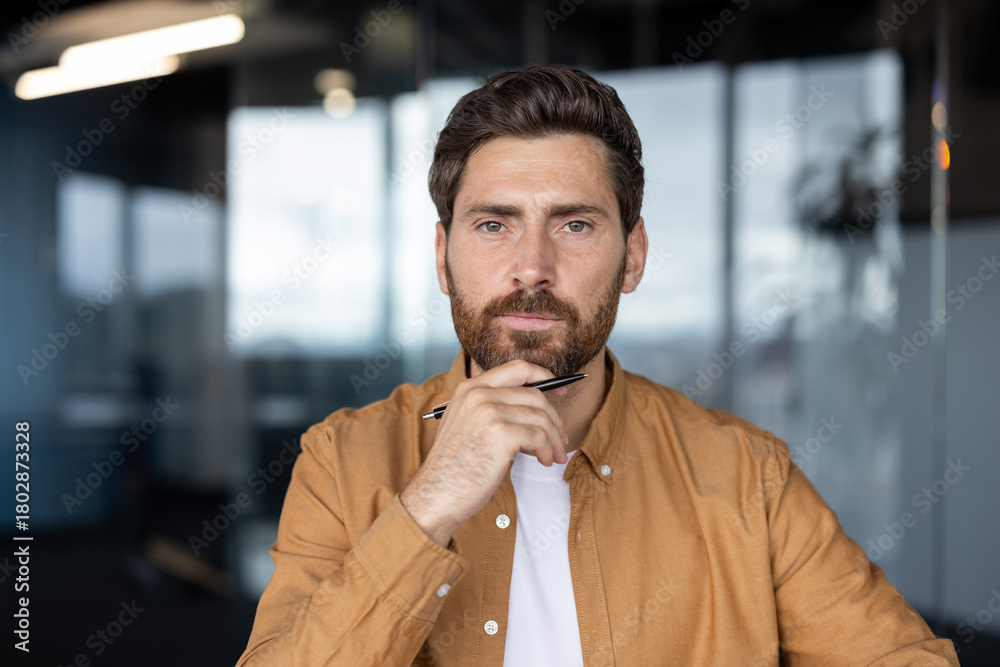 Naklejka premium Serious well-groomed professional man having an idea, looking at camera while holding a pen under his chin, planning strategy in a contemporary workplace environment