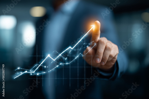A business professional is pointing at a digital growth chart that shows an upward trend. This activity is taking place in a modern office setting, emphasizing progress and success