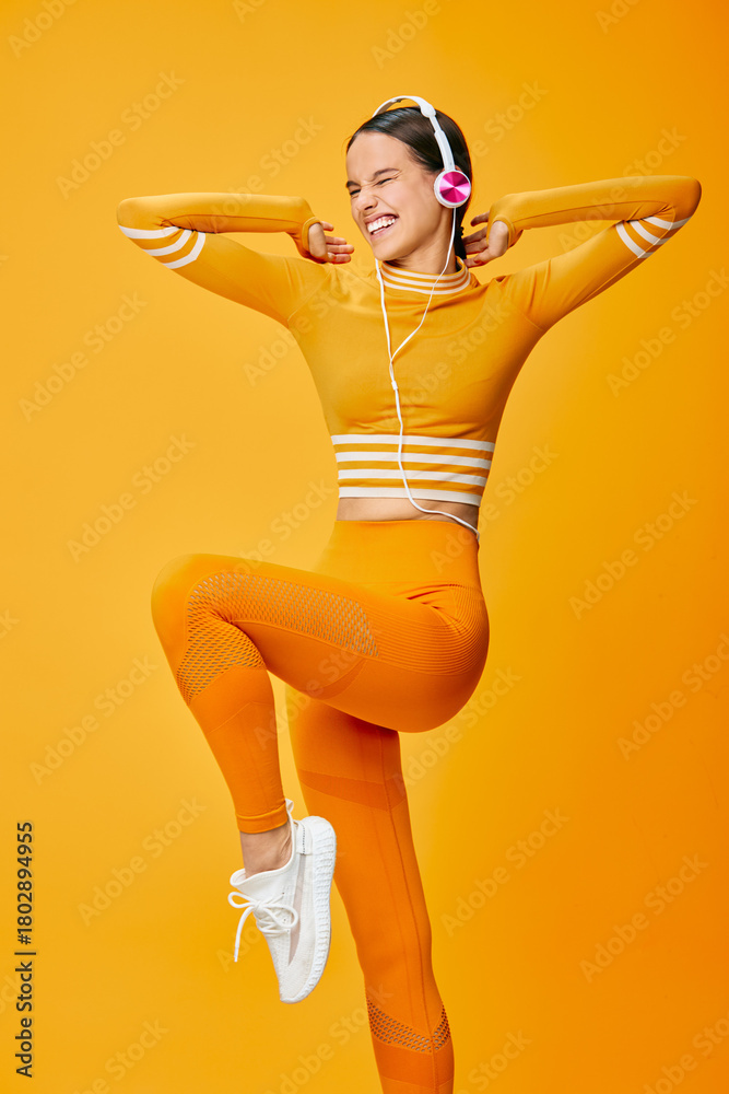 Naklejka premium Female fitness enthusiast wearing orange activewear performs a lively dance pose, exuding energy and athletic confidence in a vibrant studio backdrop. Her headphones underline a dynamic, upbeat