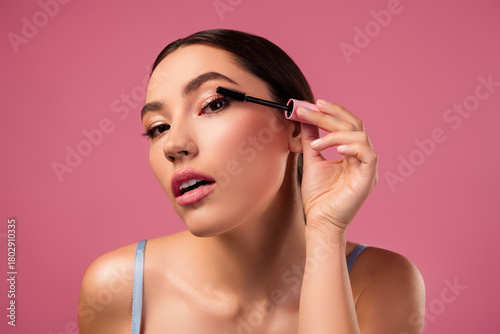 Gorgeous woman applying mascara on her eyelashes in a stylish beauty and fashion inspired photo on a pink backdrop