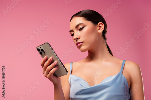 Young woman with sleek hairstyle fashionably using a modern smartphone, standing against vibrant pink background, showcasing beauty and elegance
