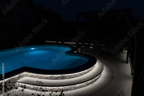 A modern outdoor pool glowing at night with vibrant LED lighting inside and around the water, creating a luxurious, serene, and atmospheric scene ideal for relaxation and nightlife visuals.