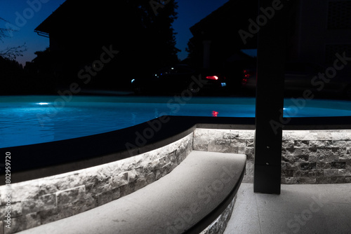A modern outdoor pool glowing at night with vibrant LED lighting inside and around the water, creating a luxurious, serene, and atmospheric scene ideal for relaxation and nightlife visuals.