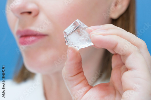 Young woman holding ice cube near her face on blue background.