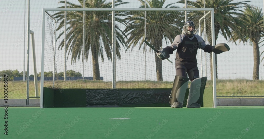 Naklejka premium Standing teen field hockey goalkeeper wearing helmet and pads, holding stick, guarding goal on turf