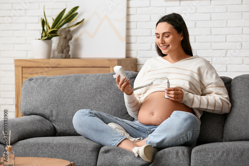 Quadro su tela Young pregnant woman with fetal doppler listening to baby's heartbeat on sofa at