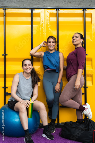 Diverse group of latin hispanic women smiling, wearing activewear, and posing against a yellow wall after a fitness session