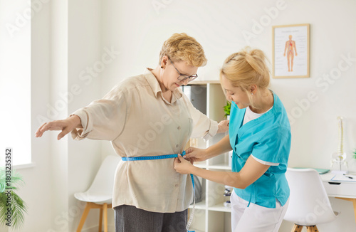 Young female healthcare worker in uniform uses measuring tape to check waist circumference of older woman patient in modern clinic or doctor office during check-up. Dietetics, weight loss concept