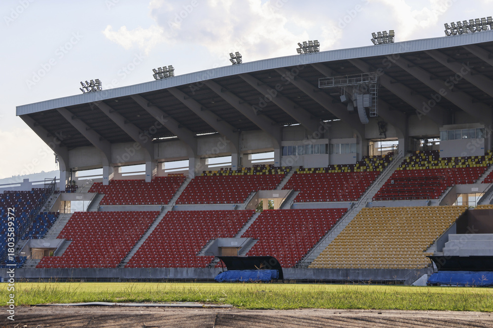 Obraz premium Quiet, empty stadium showing colorful red, blue, and yellow seats under large roof. view from green athletic field captures grandstand with feeling of anticipation