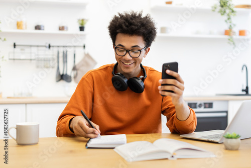 A young man with glasses smiles while looking at his phone and writing in a notebook at a table with a laptop and coffee cup.