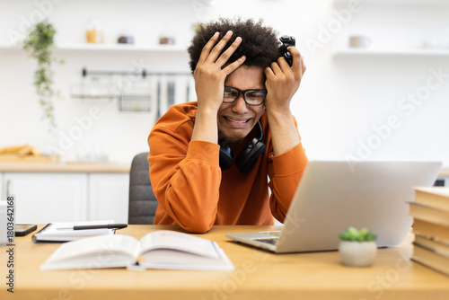 Foto A young man is visibly upset, holding a game controller, and looking at a laptop screen