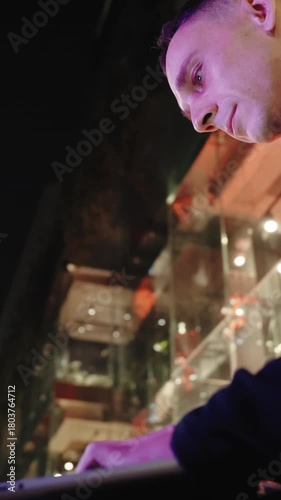 vertical close-up of a handsome Caucasian man typing on a phone, concentrating while texting under the neon glow of a city at night.