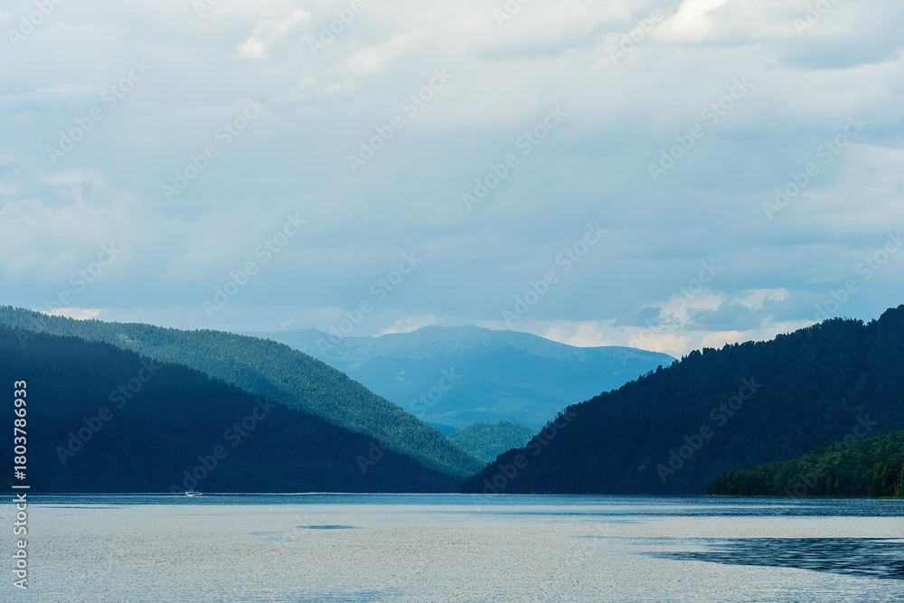 Obraz premium A picturesque view of a calm lake surrounded by forested mountains under a cloudy sky. A single boat traverses the water